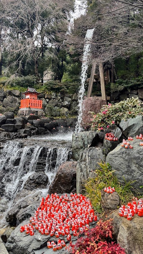 Collection of red Daruma dolls displayed beside a small waterfall and shrine at Otagi Nenbutsuji Temple in Kyoto, Japan.