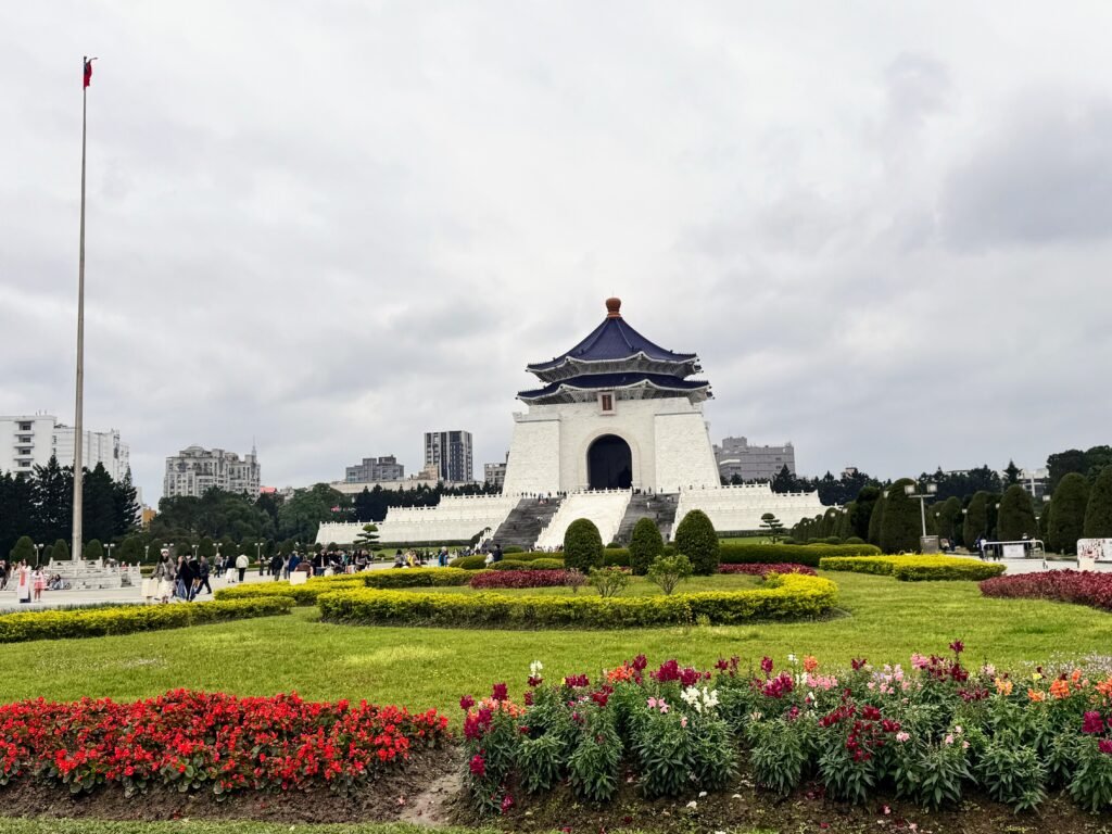 Wide view of Chiang Kai-shek Memorial Hall in Taipei, Taiwan, featuring colorful flower beds, manicured gardens, and visitors walking across Liberty Square under a cloudy sky.