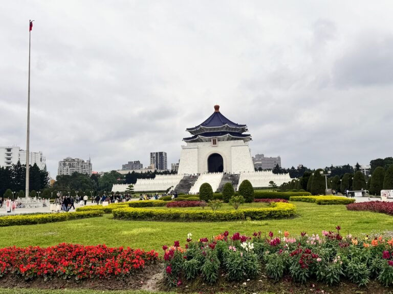Wide view of Chiang Kai-shek Memorial Hall in Taipei, Taiwan, featuring colorful flower beds, manicured gardens, and visitors walking across Liberty Square under a cloudy sky.