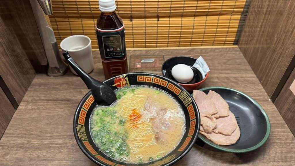 Bowl of Ichiran tonkotsu ramen topped with green onions and spicy red sauce, served with extra pork slices, a soft-boiled egg, and bottled tea in a private dining booth in Japan.